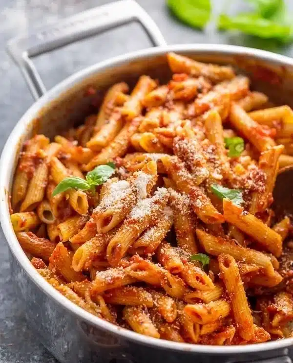Penne pasta being added to a pan of tomato sauce with basil and parmesan cheese.